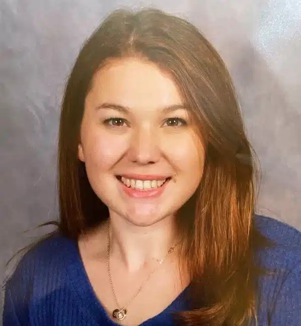 A woman with long brown hair smiles at the camera. Clad in a blue sweater, she radiates warmth, her heart pendant glinting softly. Her serene expression mirrors the calm and positivity often nurtured through pulmonary rehab against a plain, light-colored backdrop.