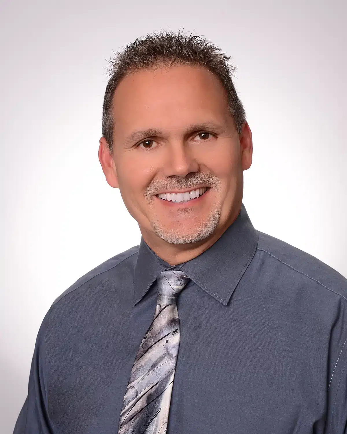 A man with short hair and a beard smiles at the camera, exuding confidence. He is wearing a gray dress shirt with a shiny tie featuring abstract patterns, perhaps ready for a day at his pulmonary rehab session. The background is plain white, focusing all the attention on him.