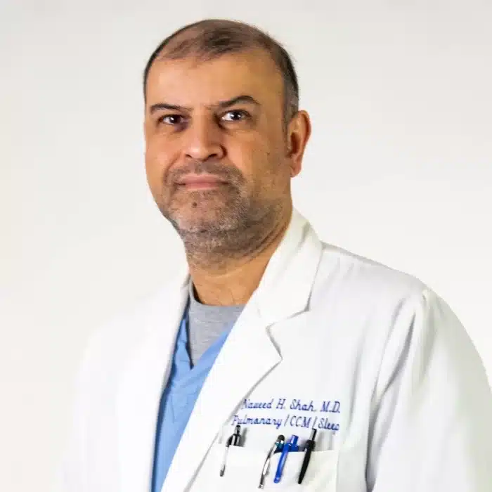 A person with short hair and facial hair, wearing a white lab coat over a blue shirt with stitched text on the left side, stands in front of a plain, light-colored background. They radiate confidence and knowledge, embodying the spirit of pulmonary rehab expertise.