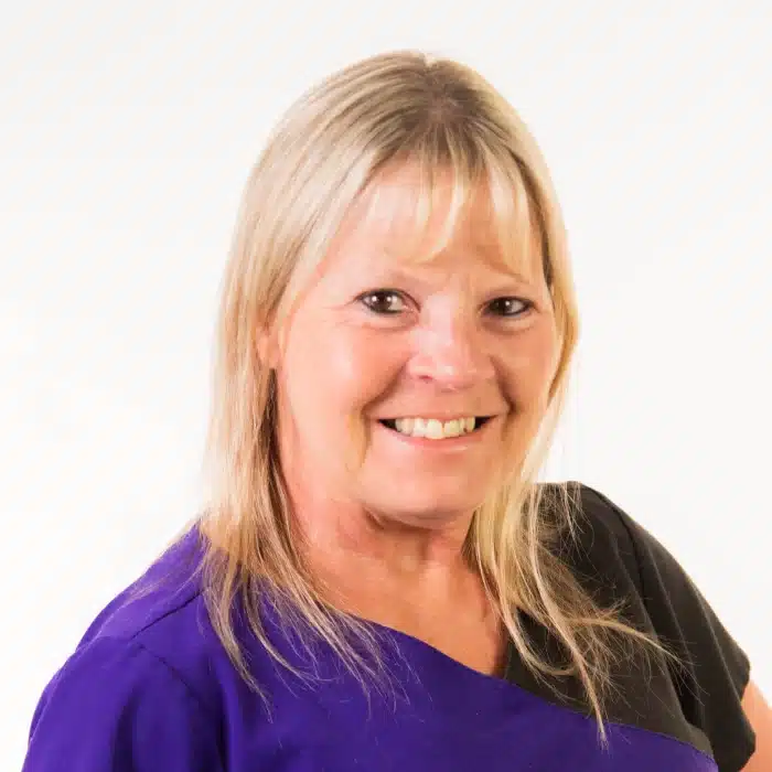 A smiling woman with long blonde hair, wearing a purple and black top, stands confidently against a white background, embodying the positive spirit of pulmonary rehab.