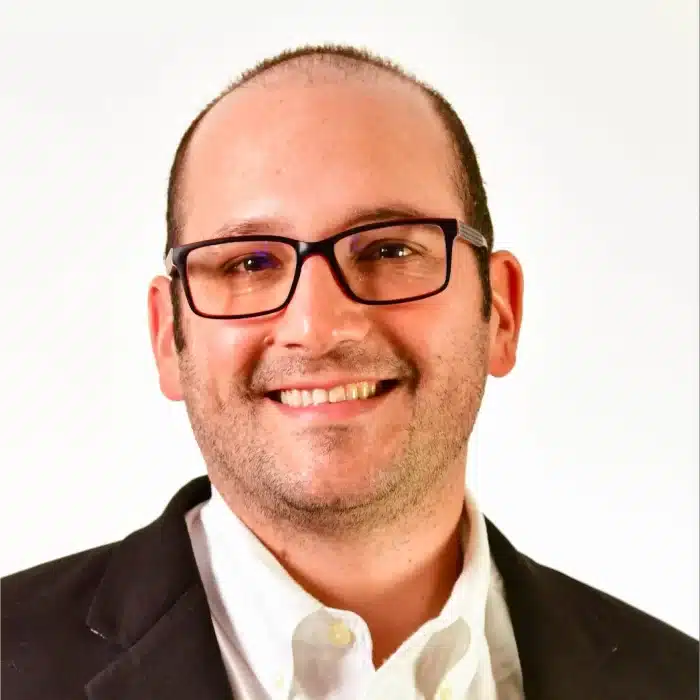 A man with glasses, short hair, and a beard, wearing a white shirt and dark blazer, smiles at the camera against a plain white background, embodying the positivity of pulmonary rehab.