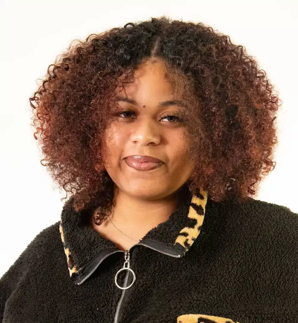 A person with curly hair and a warm smile is wearing a black fleece jacket with animal print accents, looking as if they're ready for their next pulmonary rehab session. The background is plain white, highlighting their confident presence.