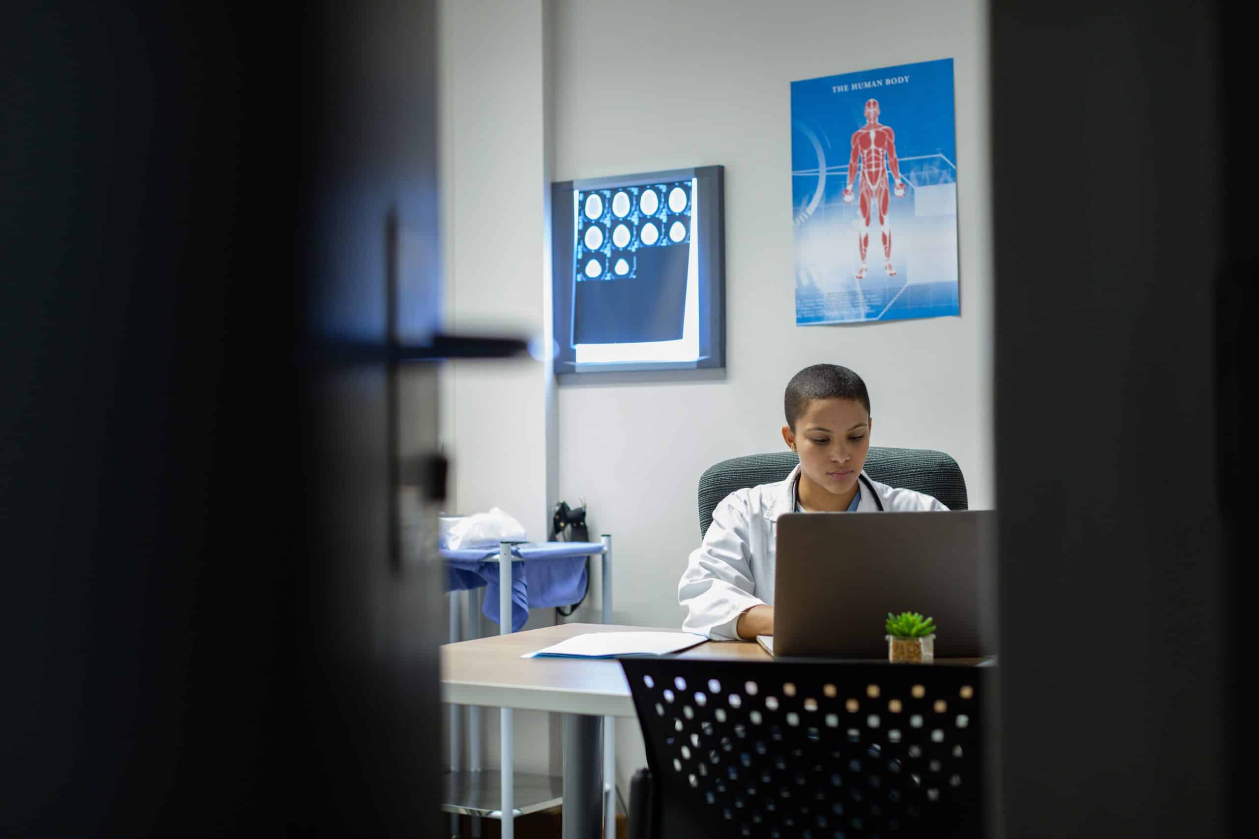 A medical professional sits at a desk focused on a laptop, researching pulmonary rehab. The office features a blue anatomy poster and illuminated brain scans on the wall. A small plant is on the desk. The scene is viewed through a partially open door, offering a glimpse into this diligent workspace.