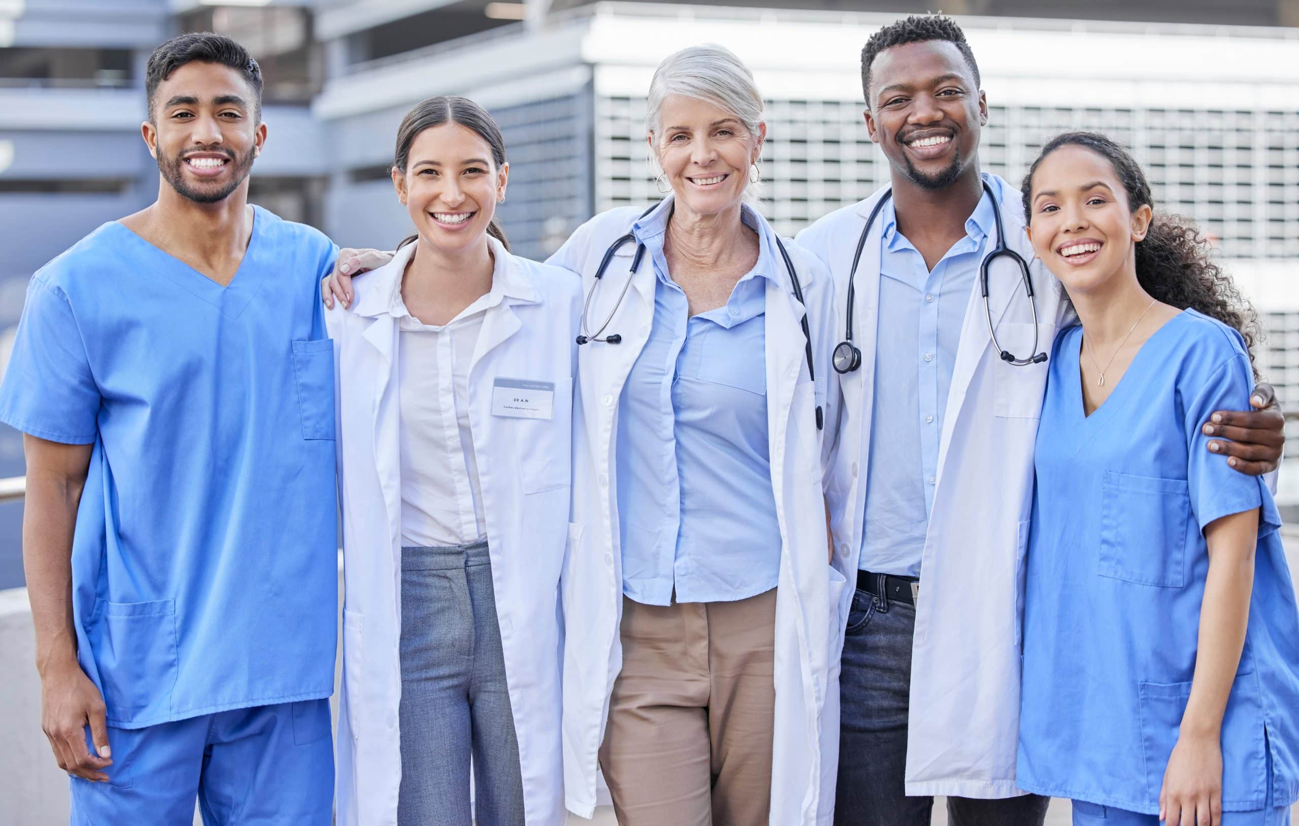 A diverse group of five healthcare professionals, smiling and wearing scrubs and lab coats, stands closely together outside a modern building. With stethoscopes around their necks, they appear ready for a day focused on pulmonary rehab, showcasing their commitment to patient care.