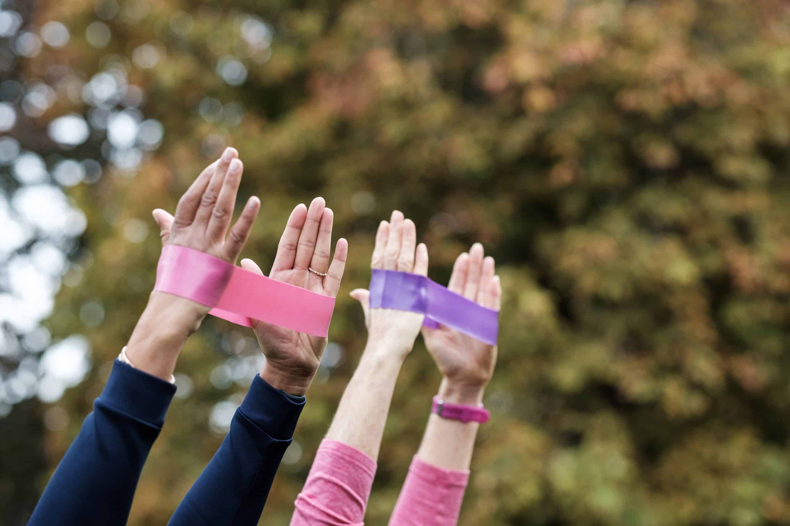 Three individuals raise their hands, each with a different colored resistance band (pink and purple) wrapped around their wrists, as part of an engaging pulmonary rehab session. The background features blurred greenery.