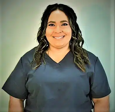 A person with long, wavy hair is smiling while wearing a dark scrub top, embodying the warmth and care often found in pulmonary rehab. The background is neutral and softly lit, enhancing the serene atmosphere.