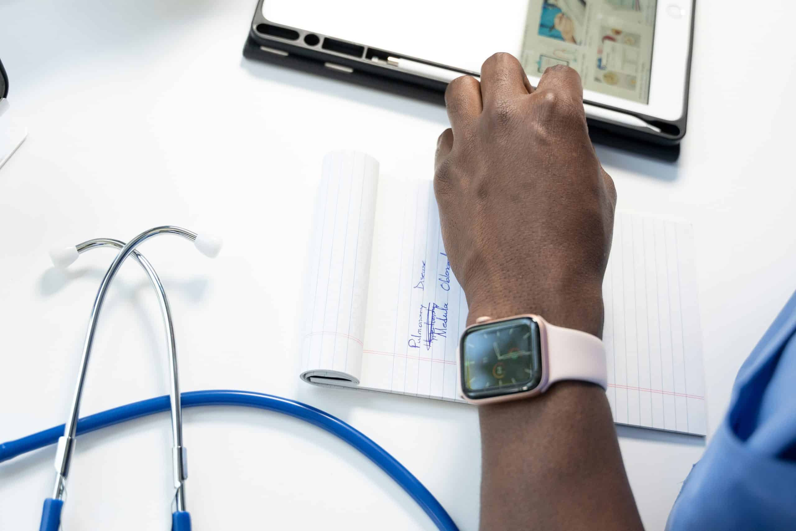 A close-up of a person writing on a notepad during pulmonary rehab. The individual, in a blue shirt and wearing a smartwatch with a white band, is surrounded by essential tools—a stethoscope and an electronic tablet on the pristine white table nearby.