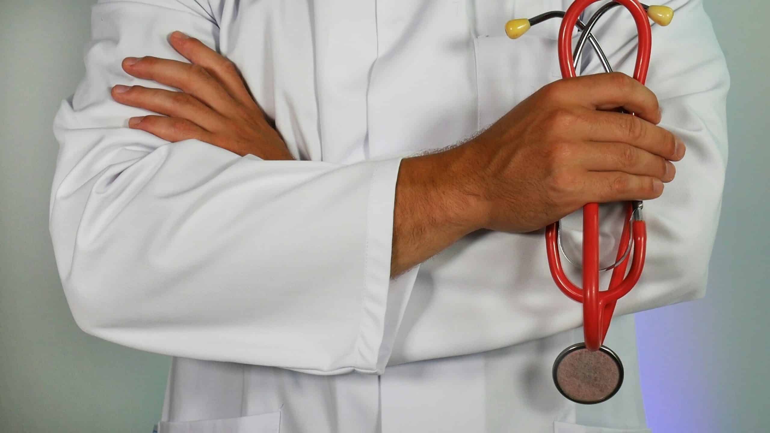 A person in a white lab coat with arms crossed, holding a red stethoscope, exudes expertise in the field of pulmonary rehab. The background is plain and out of focus.