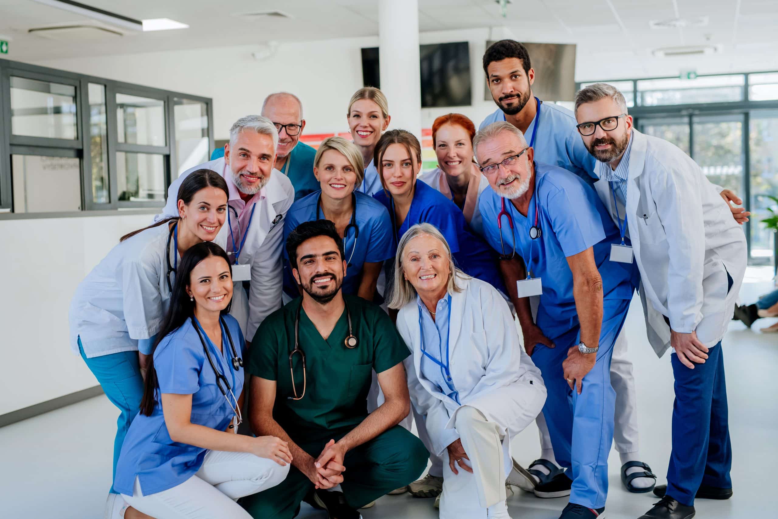 A group of medical professionals, specializing in pulmonary rehab, including doctors and nurses, are smiling and posing together in a well-lit hospital or clinic setting. They are wearing a mix of blue and green scrubs and white lab coats.