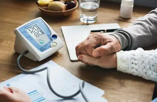 Two elderly people holding hands at a table with a blood pressure monitor displaying 120/80 mmHg. A document with charts on pulmonary rehab, a pen, and a bowl of fruit are also visible.