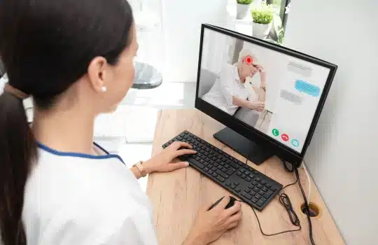 A woman is engaged in a video call on her desktop computer, connecting with an elderly man who appears to be discussing his progress with pulmonary rehab. Text messages are visible on the screen, adding a layer of interaction in the cozy setting of her home office.