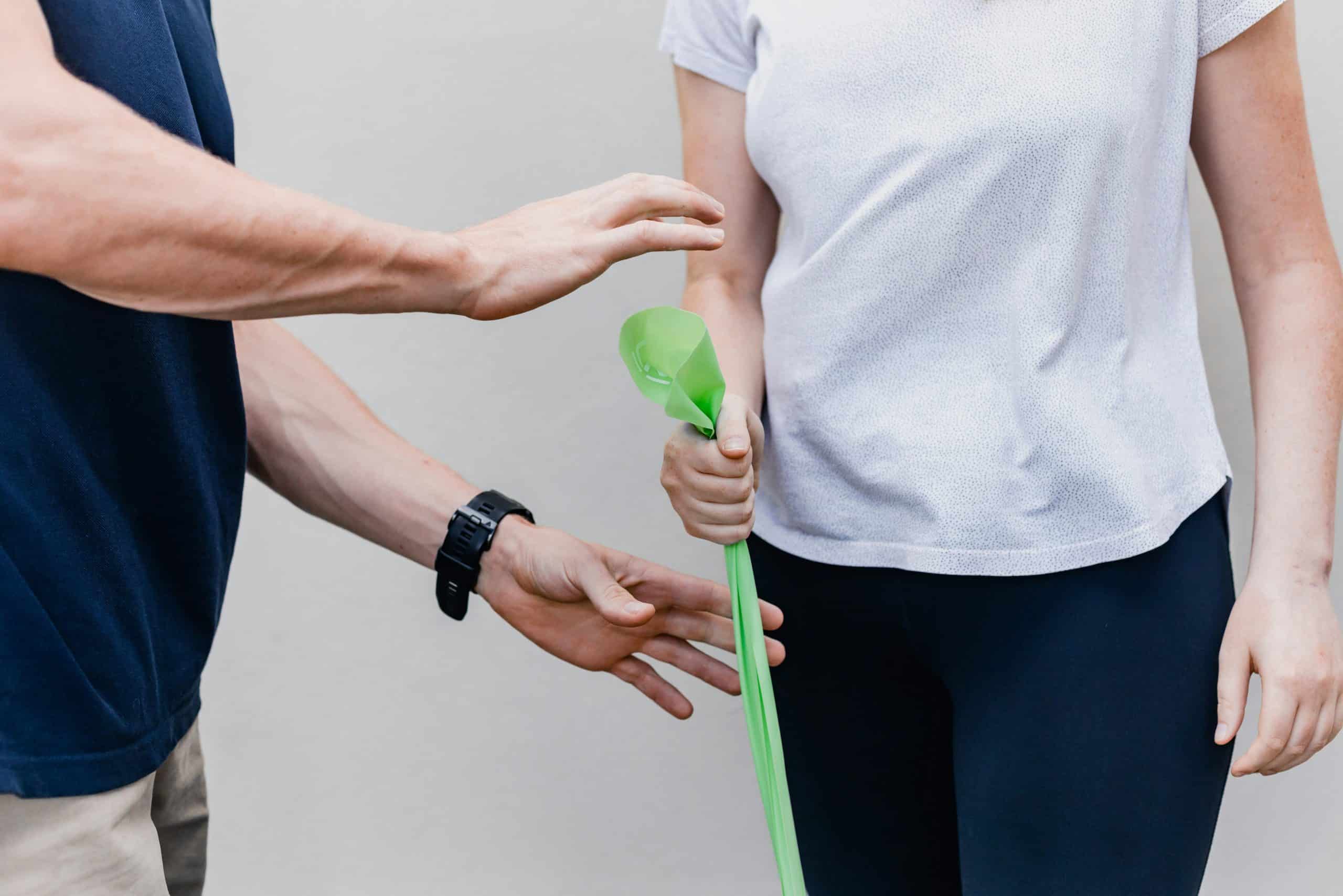 A person wearing a white shirt is holding a green resistance band attached to a wall, demonstrating an exercise often used in pulmonary rehab. Another person, in a dark shirt, stands nearby, gesturing towards the band.