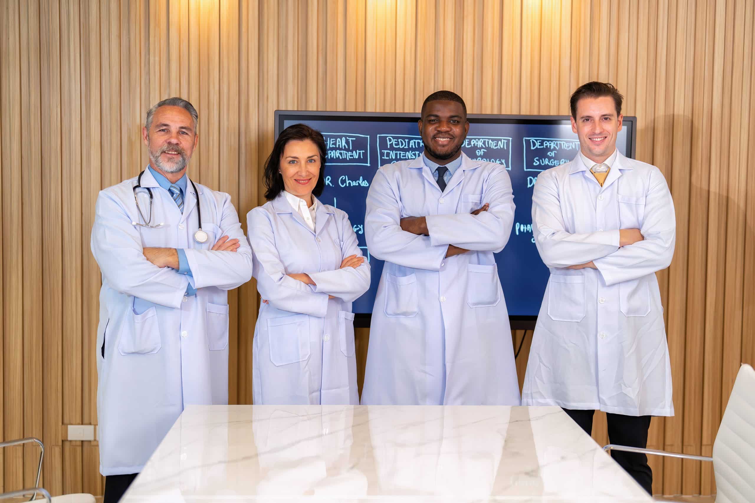 Four medical professionals in lab coats stand with arms crossed in a wood-paneled room. They are in front of a chalkboard displaying pulmonary rehab and other medical departments. A table is partially visible in the foreground.