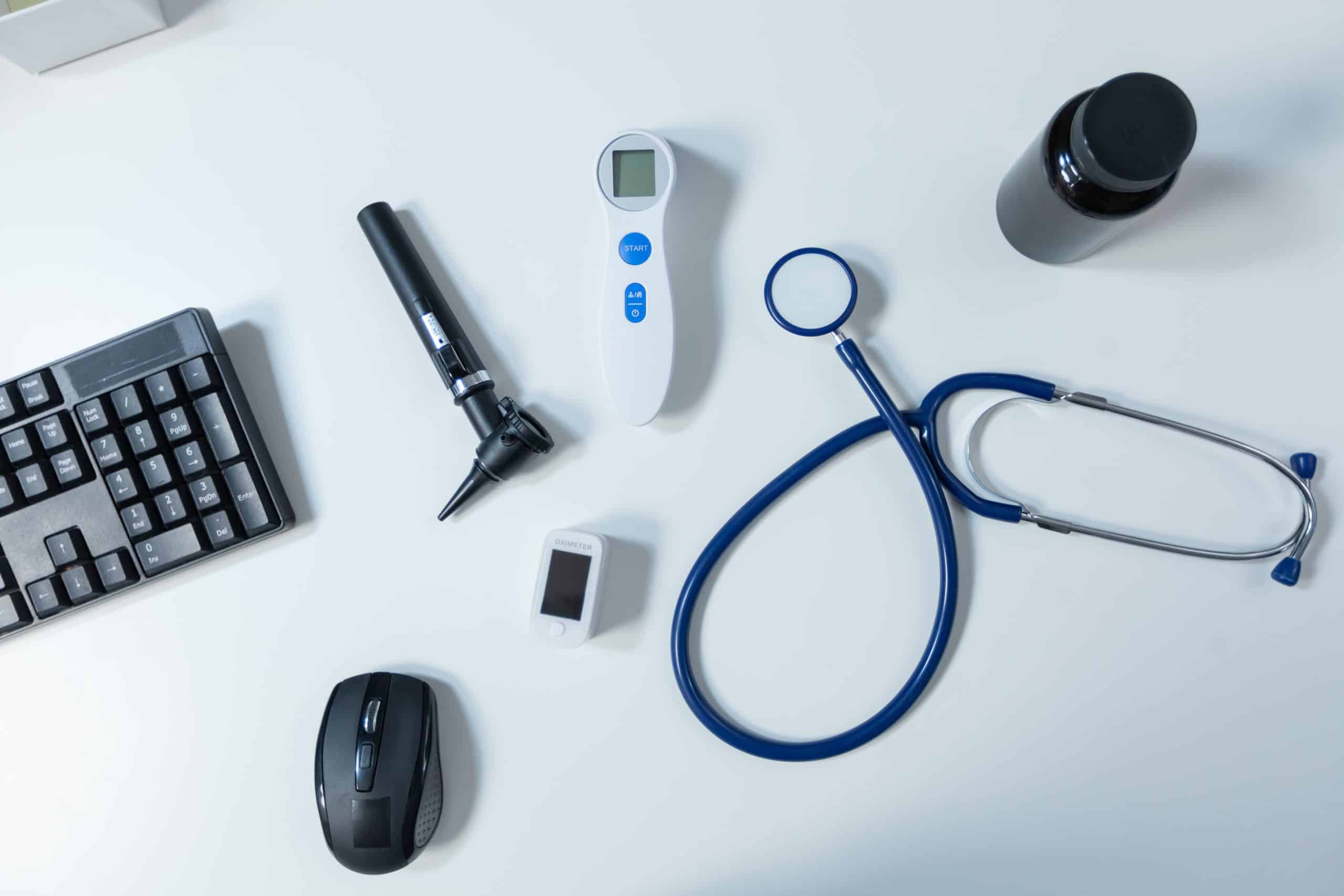 Medical tools for pulmonary rehab are on a table, including a stethoscope, digital thermometer, otoscope, and pulse oximeter. Also visible are a computer keyboard, a mouse, and a dark medicine bottle, all set against a white surface.