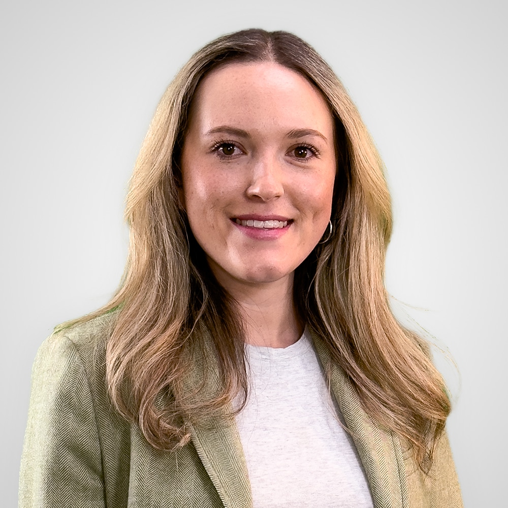 Smiling woman with long, light brown hair wearing a light green blazer and white top, standing in front of a plain, light gray background.