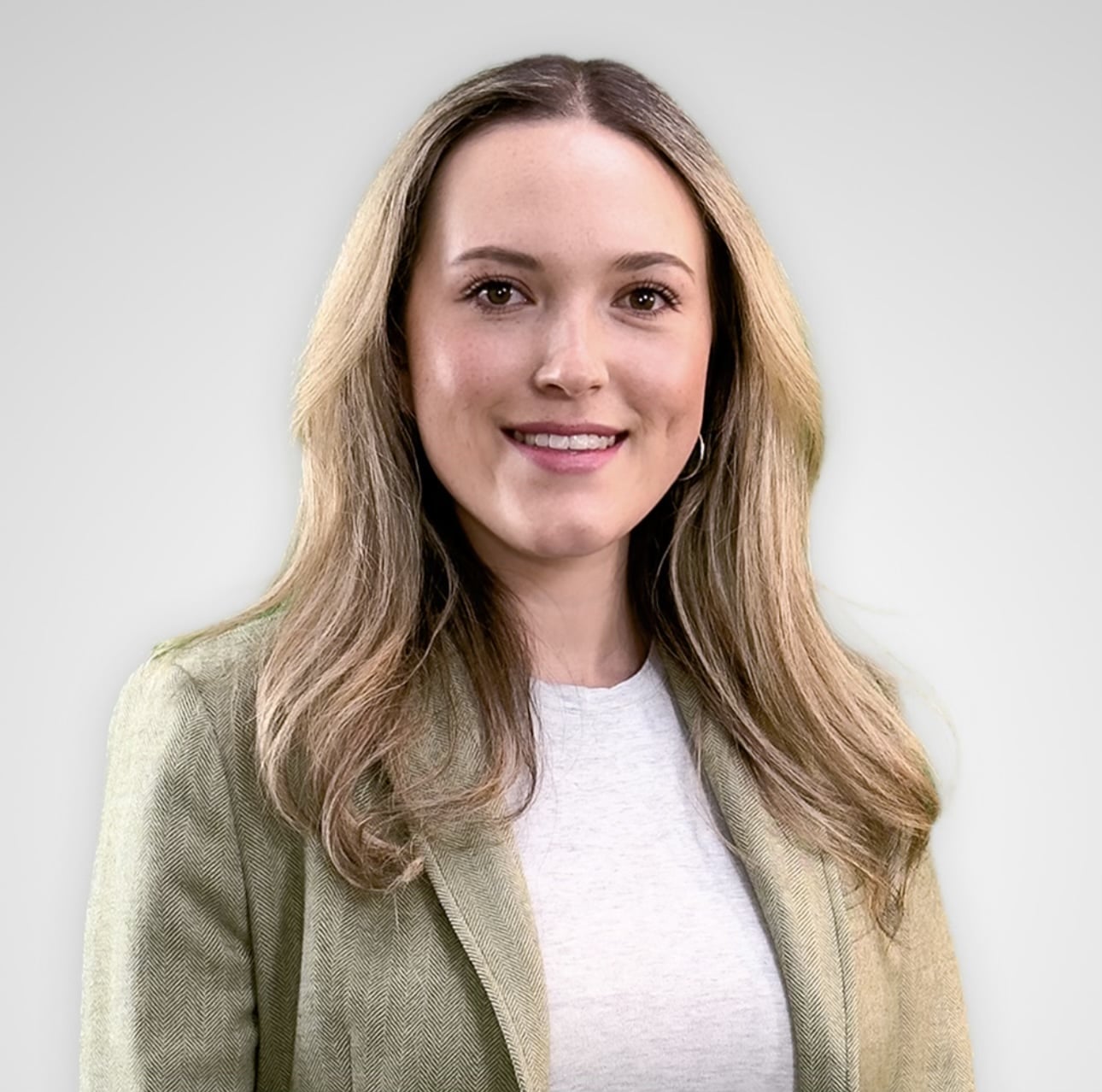 A woman with long, light brown hair smiles while wearing a light gray blazer over a white top. She stands against a neutral gray background.