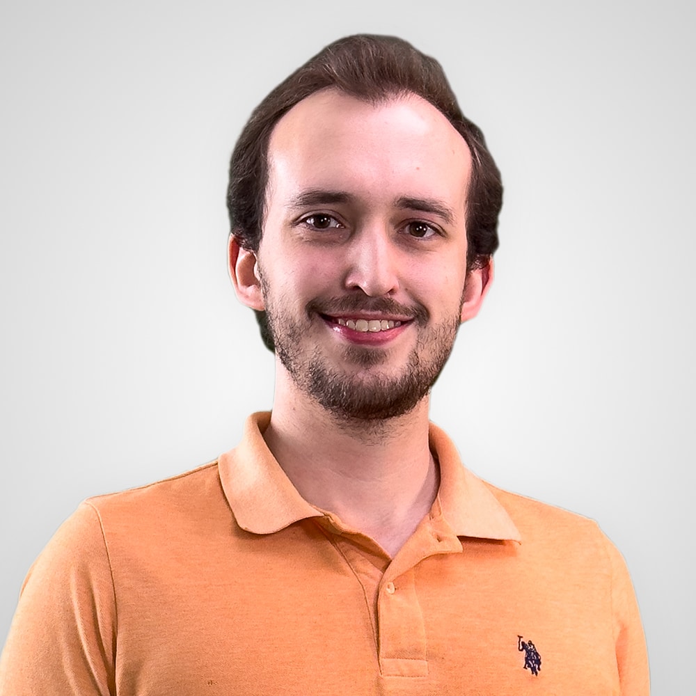 Man with short dark hair and a beard, smiling. He's wearing an orange collared shirt with a small logo on the left side against a light gray background.