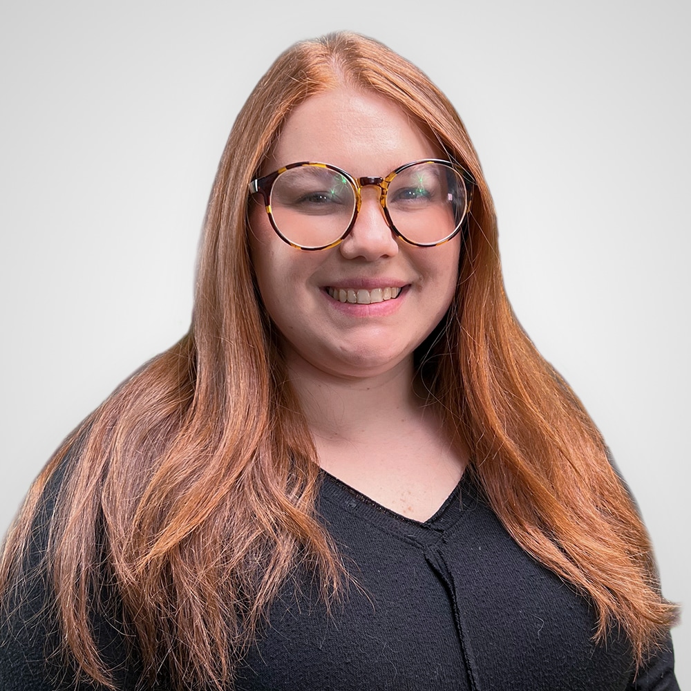 A smiling person with long, straight, reddish-brown hair and wearing large round glasses and a black top stands against a plain white background.