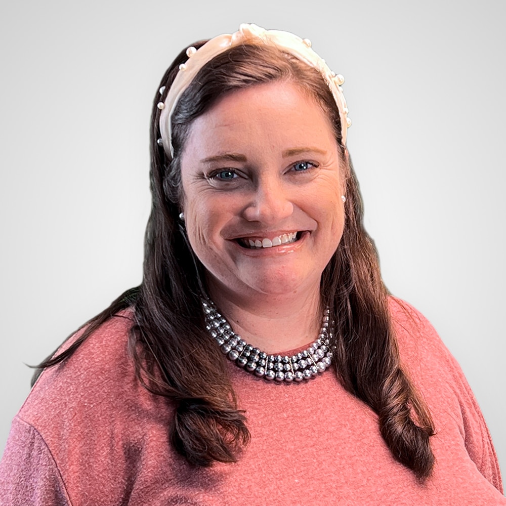 Smiling woman with long brown hair wearing a light headband with pearls, a beaded necklace, and a pink sweater, standing against a plain white background.