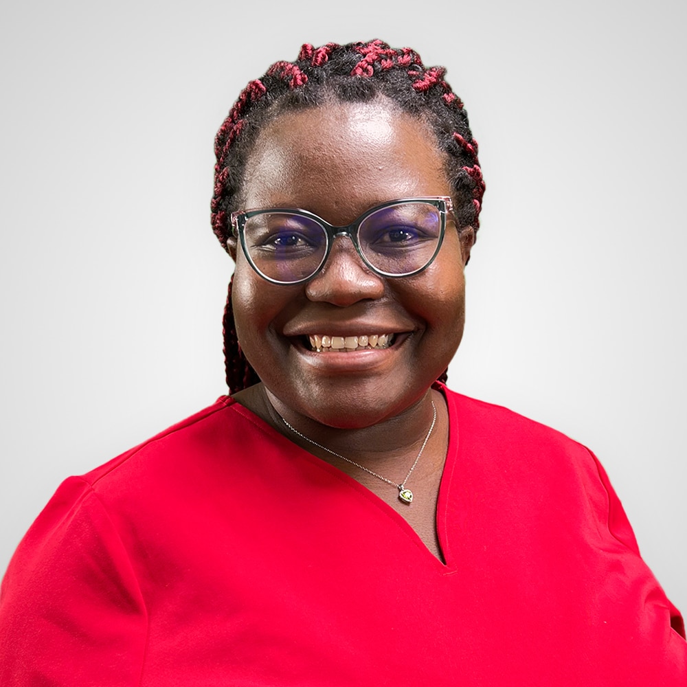 Smiling person wearing glasses and a red top with braided hair styled with red accents. The background is a plain light gray.